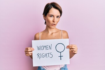 Young brunette woman with short hair holding woman rights banner clueless and confused expression. doubt concept.