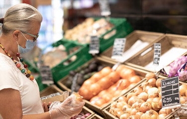 A senior retired woman white dressed  wearing surgical mask due to coronavirus shopping and choosing vegetables in a store using protective gloves