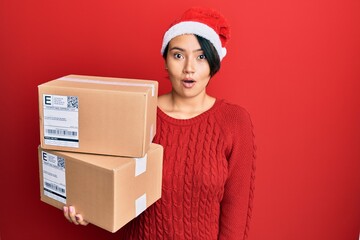 Beautiful young woman with short hair wearing christmas hat holding delivery packages scared and amazed with open mouth for surprise, disbelief face