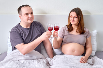 A man and a pregnant woman drink wine in bed. Husband and wife with alcohol in glasses