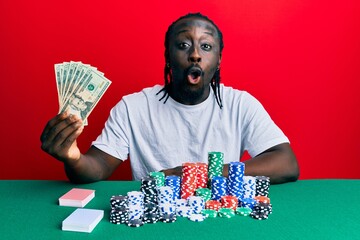 Handsome young black man playing poker holding 20 dollars banknotes scared and amazed with open mouth for surprise, disbelief face