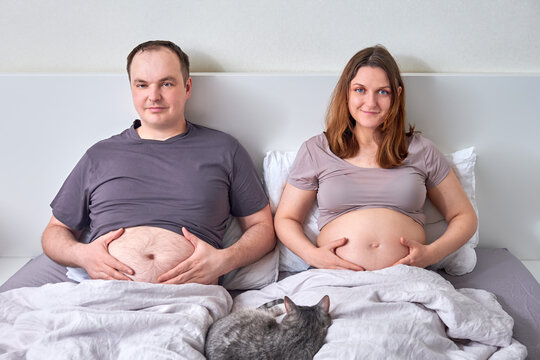 A pregnant woman and a fat man lie in bed holding their bellies, the concept of a sympathetic pregnancy