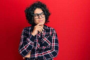 Fototapeta na wymiar Young hispanic woman with curly hair wearing casual clothes and glasses with hand on chin thinking about question, pensive expression. smiling with thoughtful face. doubt concept.
