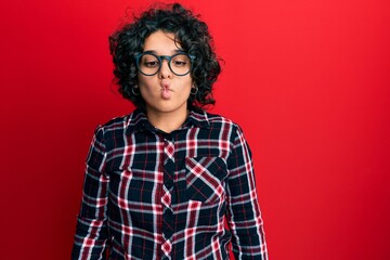 Obraz na płótnie Canvas Young hispanic woman with curly hair wearing casual clothes and glasses making fish face with lips, crazy and comical gesture. funny expression.
