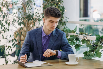 Serious young businessman in a business suit is resting in a street cafe, writes in a notebook and looks at his watch while checking the time