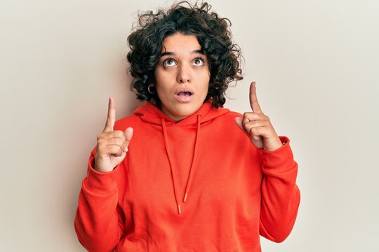 Young hispanic woman with curly hair wearing casual sweatshirt amazed and surprised looking up and pointing with fingers and raised arms.
