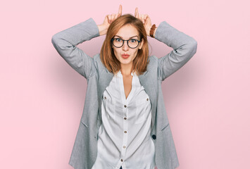 Young caucasian woman wearing business style and glasses doing funny gesture with finger over head as bull horns