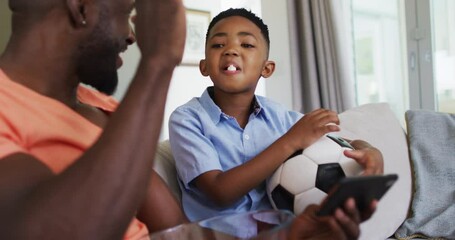 African american father and son eating popcorn and watching a football game on smartphone - Powered by Adobe