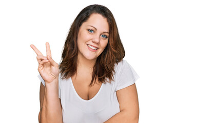 Fototapeta premium Young plus size woman wearing casual white t shirt smiling looking to the camera showing fingers doing victory sign. number two.