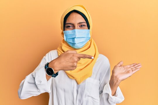 Young brunette arab woman wearing traditional islamic hijab and medical mask amazed and smiling to the camera while presenting with hand and pointing with finger.