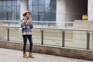 Pretty laughing caucasian woman with smartphone has good time outdoor. portrait of trendy millennial girl with blonde hair wears hat and fashionable shirt