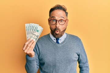 Handsome middle age man holding 20 dollars banknotes scared and amazed with open mouth for surprise, disbelief face