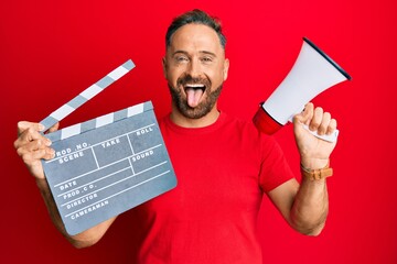 Handsome middle age man holding video film clapboard and megaphone sticking tongue out happy with...