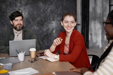 Portrait of smiling young businesswoman wearing red jacket during meeting with coworkers in office,...
