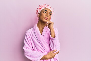 Hispanic man wearing make up wearing shower towel cap and bathrobe with hand on chin thinking about question, pensive expression. smiling with thoughtful face. doubt concept.