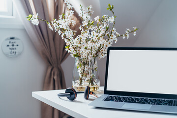 A home workplace with white screen laptop for mockup, earphones and blooming branches in a vase on the desk with room interior background. Freelance, working from home, online learning, home office.