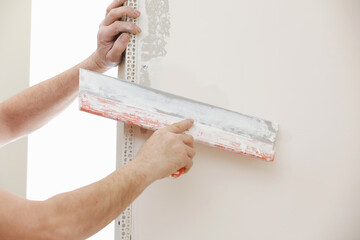 Builder in protective mask using a trowel to add plaster. Plastering wall with putty-knife, close...