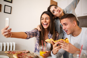 Group of young students eating pizza at the home party.Thay taking selfi with smart phone.