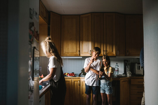 Sibling using smart phone while mother doing chores in kitchen
