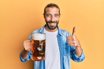 Attractive man with long hair and beard drinking a pint of beer smiling happy and positive, thumb...