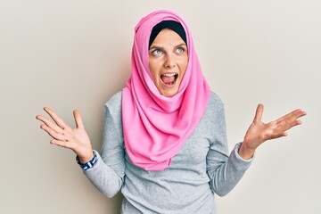 Young caucasian woman wearing traditional islamic hijab scarf crazy and mad shouting and yelling with aggressive expression and arms raised. frustration concept.