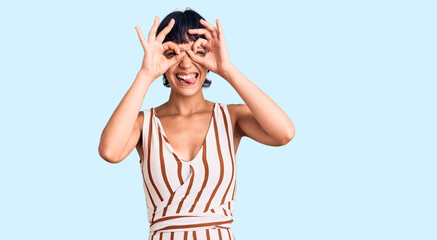 Young brunette woman with short hair wearing summer outfit doing ok gesture like binoculars sticking tongue out, eyes looking through fingers. crazy expression.