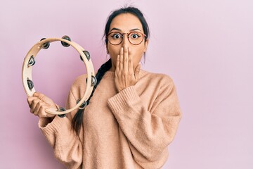 Young hispanic girl playing tambourine covering mouth with hand, shocked and afraid for mistake. surprised expression - obrazy, fototapety, plakaty