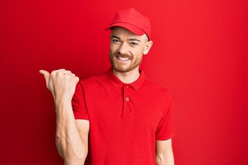 Young redhead man wearing delivery uniform and cap smiling with happy face looking and pointing to the side with thumb up.