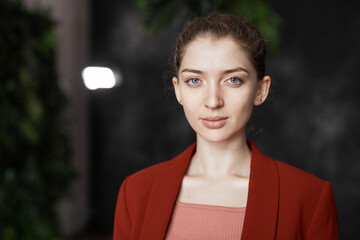 Front view portrait of successful elegant businesswoman wearing red jacket and looking at camera while posing in office, copy space