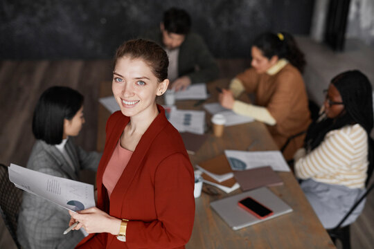 High Angle Portrait Of Successful Young Businesswoman Wearing Red Jacket And Smiling At Camera With Diverse Group Of People Meeting In Background, Copy Space