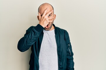 Young hispanic man wearing casual sporty jacket peeking in shock covering face and eyes with hand, looking through fingers afraid