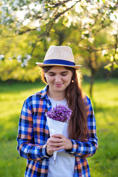 Girl holding a bouquet of lilacs in the green garden on the sunset. .Shy young girl with brown hair  in hat looked down and admires magnificent creative bouquet of flowers. Selective focus.