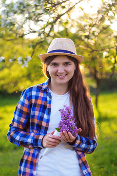 Girl holding a bouquet of lilacs in a green garden at sunset..A shy young girl with brown hair in a hat smiles and holds an original bouquet of flowers. Selective focus.