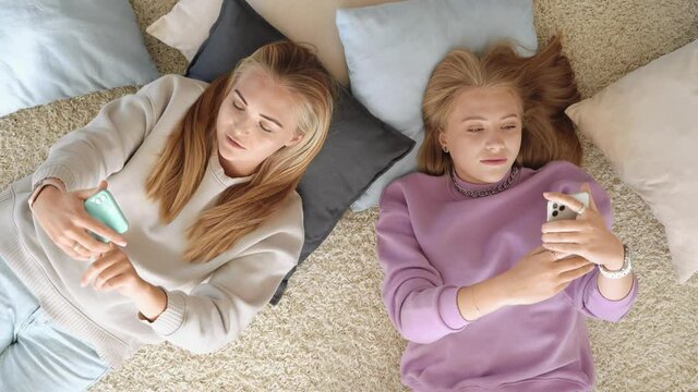 Two best girlfriends are lying on the bedroom floor among pillows, checking smartphones, relaxing and talking