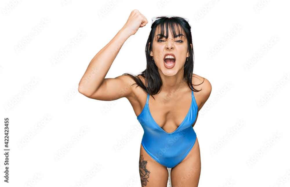 Wall mural young hispanic woman wearing swimsuit and sunglasses angry and mad raising fist frustrated and furio