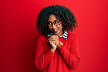 Fototapeta na wymiar Beautiful african american woman with afro hair wearing sweater and glasses laughing nervous and excited with hands on chin looking to the side