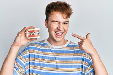 Young caucasian man holding denture winking looking at the camera with sexy expression, cheerful...
