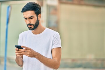 Young hispanic man with serious expression using smartphone at the city.