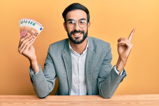Young hispanic man holding euro banknotes smiling happy pointing with hand and finger to the side