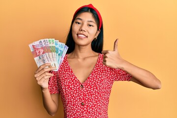 Young chinese woman holding hong kong dollars banknotes smiling happy and positive, thumb up doing excellent and approval sign