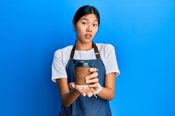 Young chinese woman wearing waiter apron holding cup of coffee clueless and confused expression....