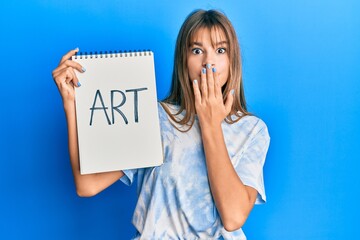 Teenager caucasian girl holding art notebook covering mouth with hand, shocked and afraid for...