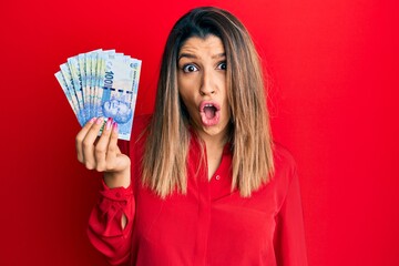 Beautiful brunette woman holding south african 100 rand banknotes scared and amazed with open mouth for surprise, disbelief face