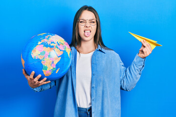 Fototapeta premium Young hispanic girl holding paper plane and world ball sticking tongue out happy with funny expression.
