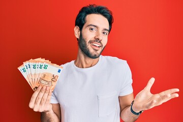 Young hispanic man holding euro banknotes celebrating achievement with happy smile and winner expression with raised hand