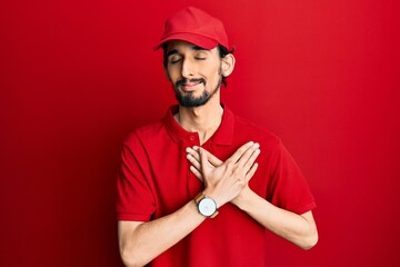 Young hispanic man wearing delivery uniform and cap smiling with hands on chest with closed eyes and grateful gesture on face. health concept.