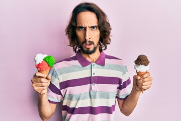 Young hispanic man holding ice cream in shock face, looking skeptical and sarcastic, surprised with open mouth