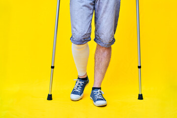 legs of a man wearing shorts and on crutches, with a bandaged leg, on yellow background