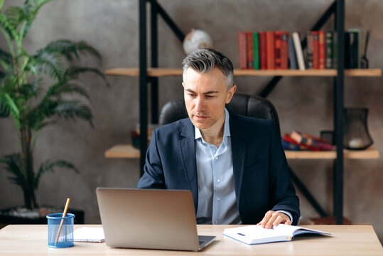 Successful Serious Adult Attractive Caucasian Business Leader Or Ceo, Sitting At Workplace In The Office In A Formal Suit, Concentrated Working In A Laptop, Browsing Internet, Analyzes Information