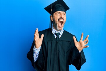 Middle age hispanic man wearing graduation cap and ceremony robe crazy and mad shouting and yelling with aggressive expression and arms raised. frustration concept.